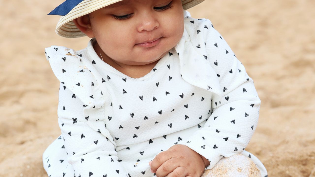Bebê sentado na areia da praia usando chapéu de proteção solar
