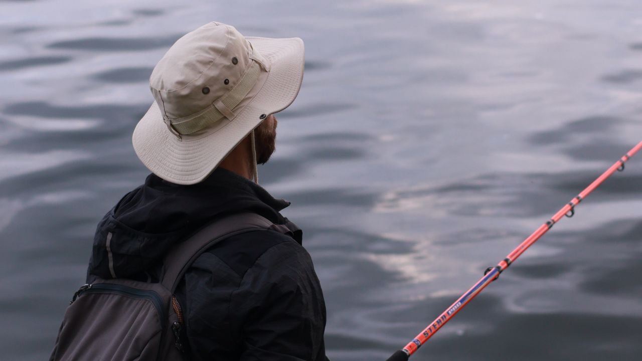Pescador com chapéu protegendo a nuca e o rosto na beira de um rio